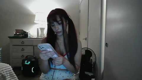 Video of a young Asian woman with long, straight red hair, wearing a pink patterned bralette and shorts, sitting in a dimly lit bedroom, focusing on her smartphone.