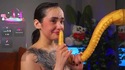 Video of a young woman with dark hair and a large tattoo on her shoulder, licking a corn cob in a dimly lit room.