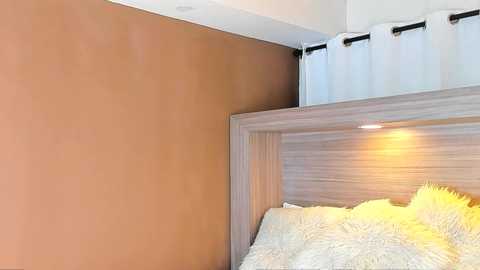 Video of a modern bedroom corner showing a light wood headboard, textured beige wall, white curtains, and a fluffy yellow pillow.