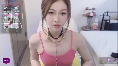 A video of an East Asian woman with light skin, brown hair, and wearing a pink tank top and gold necklace, sitting in a bedroom with a bed, flowers, and a TV.