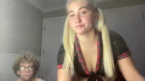 Video of a blonde woman with pigtails in a low-cut plaid shirt, smiling, and a curly-haired man with glasses, both indoors with grey walls.