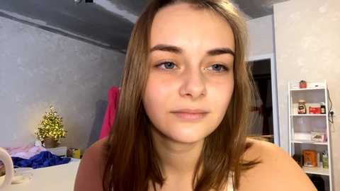 Media: Video of a young woman with long, straight brown hair and light skin, wearing a sleeveless top, in a cluttered bedroom with white walls and shelves filled with personal items.