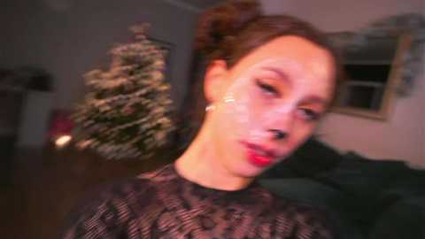 Video of a fair-skinned woman with brown hair in a bun, wearing a black lace top, with a festive background featuring a decorated Christmas tree.