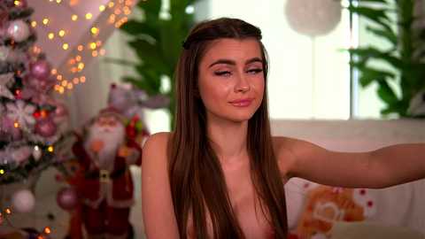 A video of a young woman with long brown hair, wearing a Santa hat, in a festive, indoor setting with a decorated Christmas tree and Santa figurines in the background.