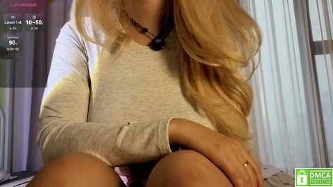 Video of a fair-skinned woman with long, wavy blonde hair, wearing a light grey long-sleeve top and shorts, sitting on a bed. The background shows a blurred room with vertical blinds.