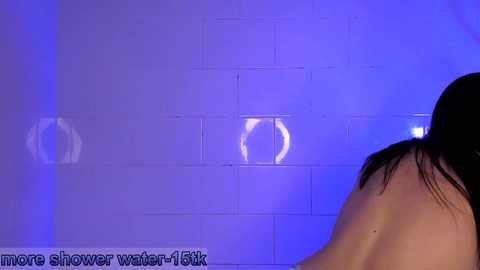 Media: A video of a person with wet, dark hair, standing in a shower with blue-tinted water, reflected in white tiles.