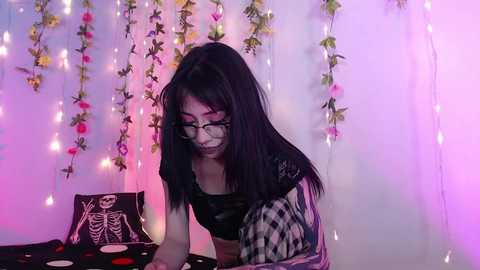 Video of a young Asian woman with long black hair, glasses, and a black crop top, sitting on a bed with a skull-patterned pillow and fairy lights in the background.