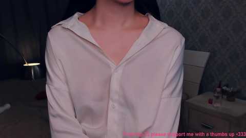 Video of a woman wearing a see-through white blouse, revealing her collarbone and cleavage, in a dimly-lit bedroom with grey wallpaper, a bedside table, and a lamp.