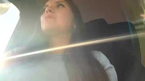 A video captures a woman with long, dark hair in a car, looking upwards through the windshield. She wears a black jacket and has a contemplative expression. Sunlight creates lens flare effects.