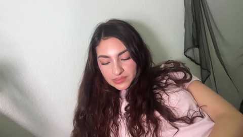 Video of a young woman with long, wavy dark hair, wearing a light pink top, standing against a plain white wall with a black mesh hanging in the background.