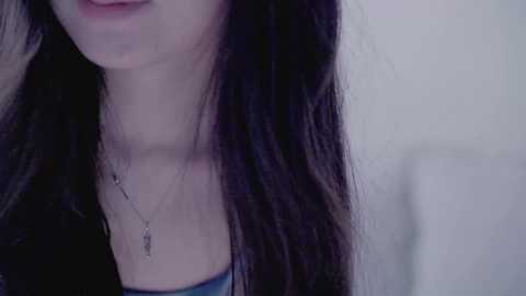 Video of a young woman with long, straight black hair, fair skin, and wearing a blue top. She has a delicate silver necklace with a small pendant. Background is out of focus, light-colored.