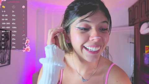 Video of a smiling young woman with light brown skin, dark hair, and pink lipstick, wearing a pink top, adjusting her hair in a bedroom with purple lighting and a mirror.