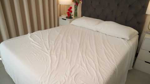Video of a neatly made bed with white sheets and pillows, gray tufted headboard, beige curtains, and a small white nightstand with a red flower vase.