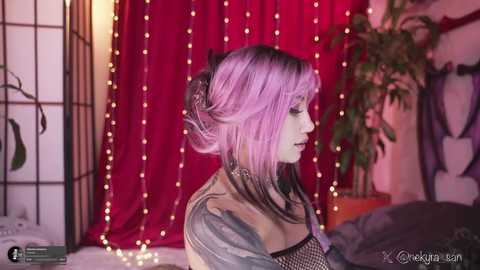 Video of a pale-skinned woman with pink and black hair, wearing a black fishnet top and a beige jacket, standing against a red curtain backdrop with fairy lights and a green plant.
