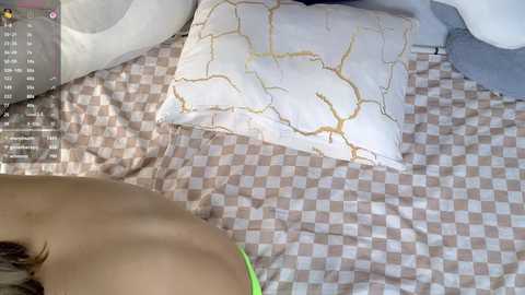 Video of a woman lying on a bed with a checkered beige and pink bedspread, wearing a lime green bikini top, with a white pillow featuring a cracked gold design beside her.