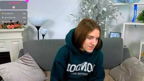 Video of a young woman with straight brown hair, wearing a dark green hoodie, sitting on a gray couch in a modern living room.