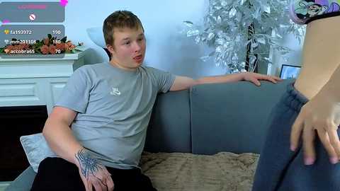 Video of a young, light-skinned man with short brown hair wearing a gray t-shirt, sitting on a gray couch, with a hand on his crotch, while a woman in jeans stands in front of him.