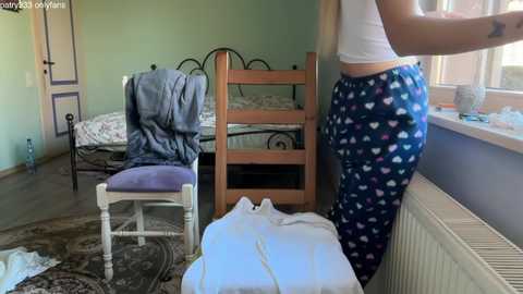 Media: Video of a messy bedroom with a young woman in heart-pattern pajama pants and a white top standing by a window. Bed, chair, and clothes scattered on the floor.
