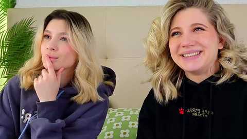 Video of a young woman with shoulder-length, wavy blonde hair, fair skin, and a broad smile, wearing a black hoodie with a red maple leaf patch, seated on a beige couch with green floral pillows.