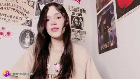 Video of a young Asian woman with long black hair, fair skin, and a light beige t-shirt. She stands against a wall adorned with vintage posters, including one featuring a man with a guitar.