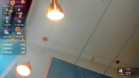 Video of a modern, dimly-lit room with a white ceiling, two hanging gold pendant lights, and a blue wall. An overlay displays a virtual reality game interface with player statistics.