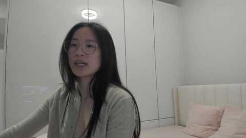 Video of an East Asian woman with long black hair, wearing glasses and a light gray cardigan, sitting on a bed in a minimalist, white-walled bedroom with a cushioned headboard.
