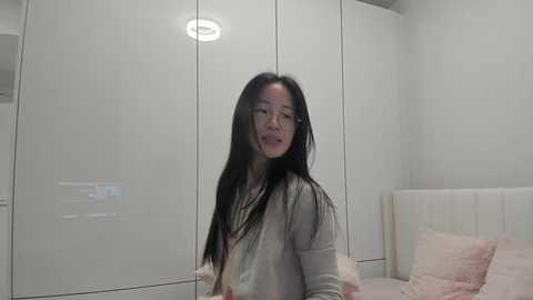Video of an Asian woman with long black hair and glasses, wearing a beige cardigan, standing in a minimalist, white bedroom with a bed and built-in wardrobe.