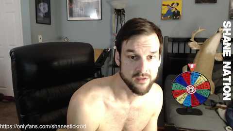 Video of a bearded, shirtless white man with short brown hair, sitting in a living room with a black leather chair, deer antlers, and colorful dartboard in the background.