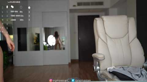 Video of a modern, minimalist living room with a large white leather chair and a reflection of a woman in a mirror. The background features white walls, wooden flooring, and a potted plant.