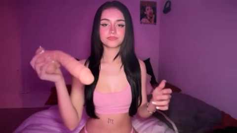 A video of a young woman with long black hair and pale skin, wearing a pink crop top, holding a prosthetic arm, in a dimly lit bedroom with purple walls.