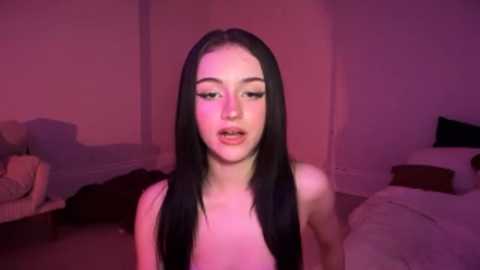 Video of a young, light-skinned woman with long black hair, wearing black lingerie, in a dimly lit, pink-hued bedroom with a bed and furniture.