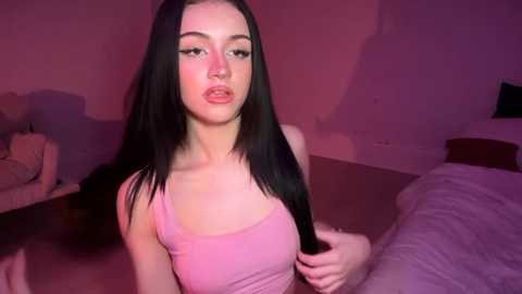 A video of a young, slender Caucasian woman with long, straight black hair and light skin, wearing a pink tank top, standing in a dimly lit bedroom with purple lighting.