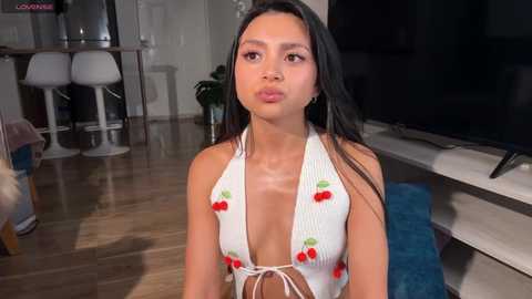 Video of a young Asian woman with long black hair, wearing a white halter top with cherry print, sitting on a blue cushion in a modern living room with wooden floors, white stools, and a flat-screen TV.