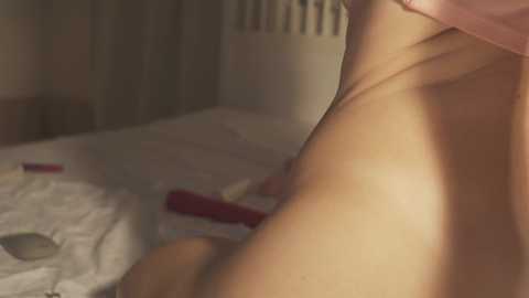 Video of a naked man, with a red vibrator on a bed, showing a close-up of his muscular back and arm. The background features beige curtains and a partially visible white radiator.