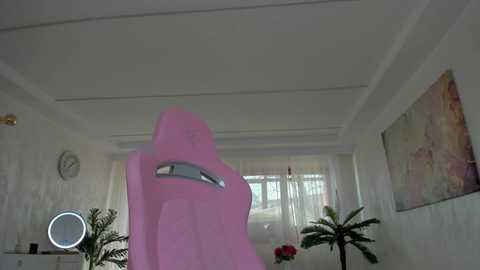 A video of a pink gaming chair in a minimalist, white-walled room with a large window, palm tree, and abstract art on the wall.
