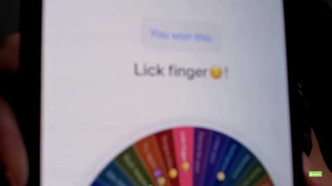 A blurred video of a smartphone screen displaying a white background with a \"Lick Finger\" notification. The screen is partially covered by a colorful, rainbow-patterned finger, suggesting a playful, interactive element.