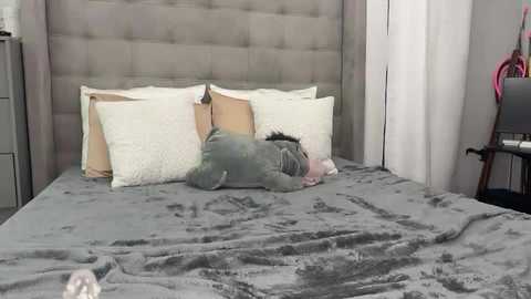 Video of a man in a gray sweatshirt and white face mask sleeping on a gray, tufted headboard bed, surrounded by white pillows, in a modern, minimalist bedroom with a pink chair and gray dresser.
