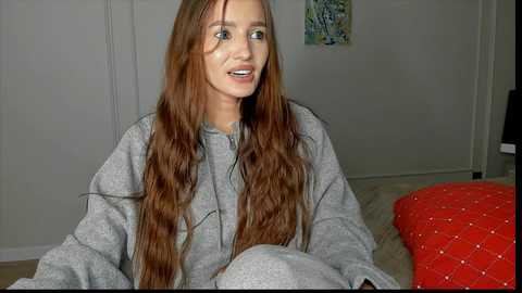 Video of a young Caucasian woman with long, wavy auburn hair, wearing a grey hoodie and pants, sitting on a bed with a red quilted pillow. Background features a beige wall with a colorful abstract painting.