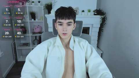 Video of a young Asian man in a white lab coat, sitting in a modern living room with white shelves, plants, and a fireplace.