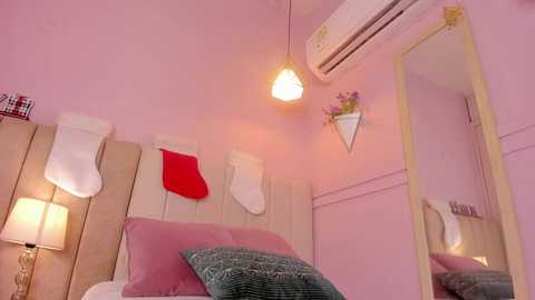 Video of a cozy, pink-themed bedroom with a wooden headboard, white socks, a red sock, and a green pillow on the bed. A mirror, air conditioner, and a flower arrangement adorn the walls.
