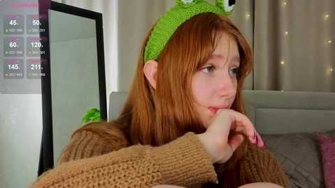 Video of a young red-haired woman wearing a green frog hat, looking contemplative with her chin resting on her hand, in a cozy bedroom.