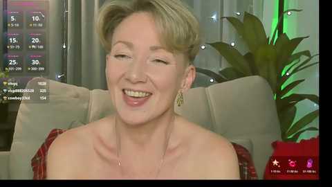 Video of a smiling, fair-skinned, short-haired woman with a red plaid shirt, seated on a beige couch in a living room with a green plant.