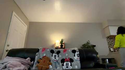 Video of a cozy living room with a plush, Mickey Mouse-themed blanket on a dark leather couch, a potted plant on a small table, and a woman standing in the background.