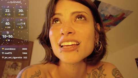 Media: Video of a tattooed, light-skinned woman with shoulder-length brown hair, multiple facial piercings, and hoop earrings, smiling. Background shows a digital screen with weather details.