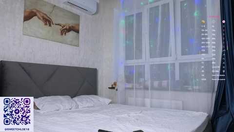 Video of a minimalist bedroom with a grey upholstered headboard, white bed linens, and sheer curtains; a large window and an air conditioning unit on the wall.