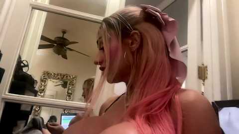 A video of a young woman with long, pink-streaked hair in a mirror, wearing a sleeveless top, inside a brightly lit room with a ceiling fan and decorative mirror.