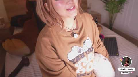 A video of a young woman with light skin and freckles, smiling in a cozy bedroom with a tan hoodie featuring a cartoon character.