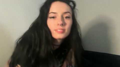 Video of a pale-skinned young woman with long, straight black hair, wearing a black shirt, and a nose ring, against a plain beige background.