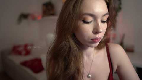 Video of a young woman with long, light brown hair, wearing a red spaghetti strap top and a silver necklace, in a softly lit, cozy living room with Christmas decorations.