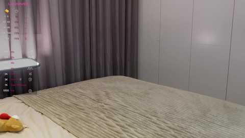 A video of a minimalist bedroom with a beige quilted bedspread, grey curtains, a white wall, and a white nightstand with a pink and yellow object on it.